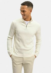 Krämfärgad kabelstickad tröja med ståkrage och dragkedja, bärs över en vit skjorta, matchad med ljusbeiga byxor.