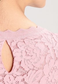 Haut en dentelle rose avec un décolleté festonné et un petit trou en forme de clé à l'arrière. Motif florale délicat sur l'ensemble du tissu.