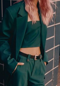 Pionowy zielony żakiet dopasowany z pasującym krótkim topem z szerokimi ramiączkami; dopasowane spodnie z czarnym paskiem i srebrną klamrą. Gładki materiał.