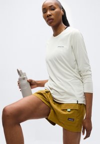 Γυναίκα με λευκό πουκάμισο μακρυμάνικο και μουσταρδί σορτς κρατά ένα γκρι παγούρι νερού, ποζάροντας με ένα γόνατο σηκωμένο μπροστά σε απλό φόντο.