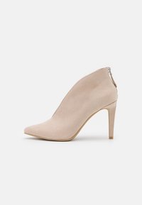Béžová semišová kotníková bota s špičatou špičkou, štíhlým vysokým podpatkem a detailem zadního zipu. Hladká textura a elegantní silueta.