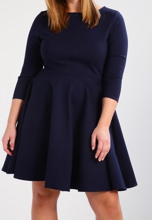 Mujer vistiendo un vestido azul marino hasta la rodilla con mangas tres cuartos, de pie sobre un fondo claro y liso.