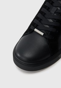 Čierne tenisky s hladkým koženým špičkou a mäkkým semišovým vrchom, s plochými čiernymi šnúrkami a kovovým logom blízko šnúrok.