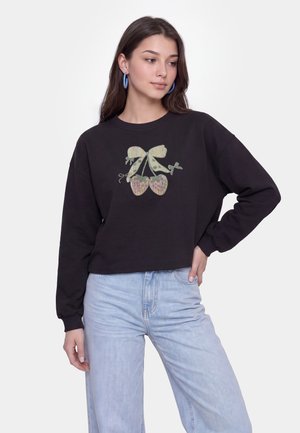 Donna con felpa nera con decorazione di ciliegie glitterate e jeans azzurri chiari, guarda di lato con la mano sui fianchi e orecchini a cerchio blu.