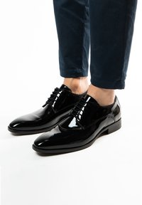 Zwarte gepatineerde leren dresschoenen met een strakke uitstraling, puntige neus en vetersluiting, gecombineerd met donkere cropped trousers.