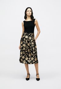 Μαύρο φόρεμα με εφαρμοστό τοπ, φούστα που ανοίγει προς τα κάτω με χρυσά ανθικά σχέδια, μήκος μέχρι τον αστράγαλο, υφασμάτινη υφή, συνδυασμένο με μαύρα τακούνια.