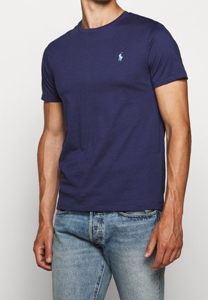 Mann trägt ein navyblaues kurzärmliges T-Shirt mit einem kleinen hellblauen Polo-Spieler-Logo auf der linken Brust und hellblauen Jeans.
