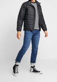 Sort dunjakke med opretstående krave, båret over en sort-hvid stribet skjorte. Et par blå jeans og sorte høj-top sneakers.