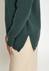 Dunkelgrüner Pullover mit weicher Textur, seitlichen Schlitzen und gerippten Bündchen, kombiniert mit hellbeigen Hosen.