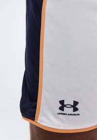 Κοντινό πλάνο ενός ατόμου που φορά λευκό και σκούρο μπλε αθλητικό σορτς με πορτοκαλί περίγραμμα και λογότυπο Under Armour κοντά στο τελείωμα.