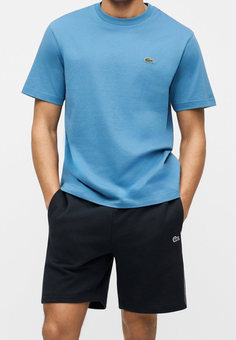 Homme portant une chemise bleue à manches courtes et un short noir, mains dans les poches, debout devant un fond blanc uni.
