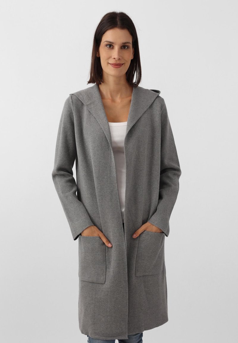 Manteau long gris avec un col châle, deux poches avant et une coupe droite, fabriqué en tissu texturé. Porté par-dessus un haut blanc.