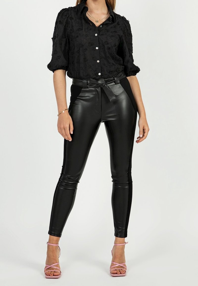 Blouse noire à texture avec des manches bouffantes, assortie à un pantalon en cuir noir taille haute avec une ceinture. Le modèle porte des talons roses à lanières.