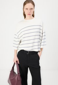 Cream striped oversized top with gray stripes, paired with black trousers. The model holds a burgundy leather shoulder bag.