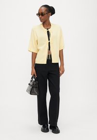 Woman wearing yellow short-sleeve cardigan with tie closures, black crop top, black wide-leg pants, black shoes, sunglasses, and holding a small black handbag.
