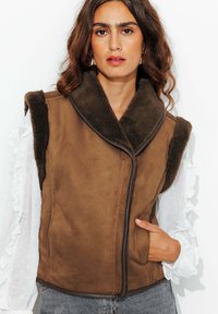 Gilet sans manches marron avec un col en fausse fourrure, présentant une texture lisse, une fermeture éclair sur le devant et des poches latérales, associé à un chemisier blanc à volants.