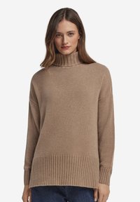 Beige stickad tröja med hög ribbad krage och ribbad fåll. Har långa ärmar och en avslappnad passform. Mjuk textur med subtil variation.