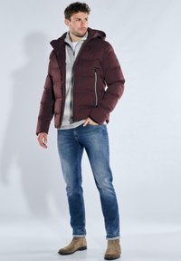Dunkelrote Steppjacke mit Kapuze, seitlichem Reißverschluss und elastischen Bündchen. Wird über einem grauen Pullover getragen und mit blauen Jeans sowie hellbraunen Stiefeln kombiniert.