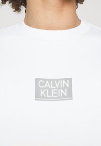 T-shirt bianca in cotone con colletto rotondo, caratterizzata da un logo rettangolare grigio che riporta la scritta "CALVIN KLEIN" in lettere in grassetto.