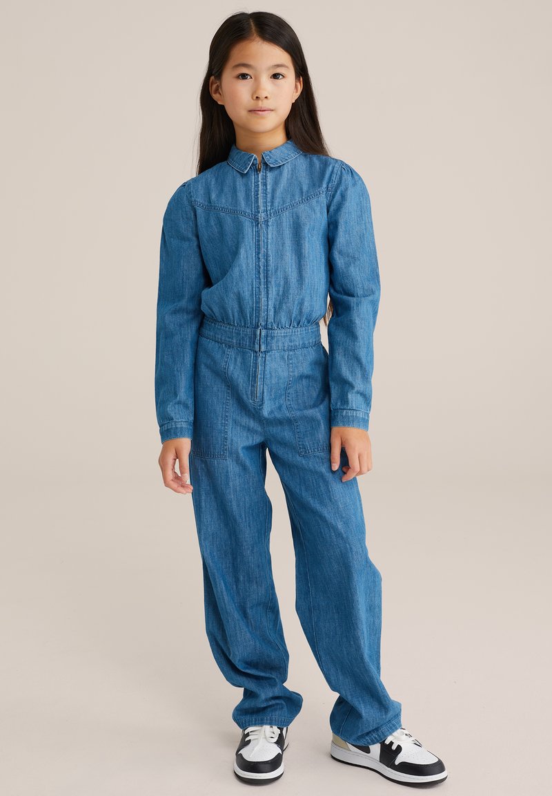 Jeanovni kombinezon z dolgimi rokavi, ovratnikom, elastičnim pasom in dvema sprednjima žepoma. Nosi se z črnimi in belimi supergami.