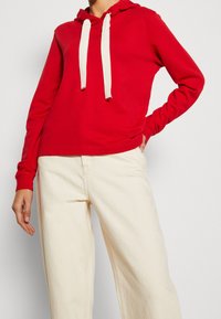 Sudadera roja con capucha y cordones blancos, hecha de material suave. Presenta mangas largas y un corte relajado, combinada con pantalones color crema.