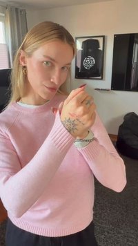 Ung kvinne med blondt hår iført en rosa genser viser tatoveringer på hånden, stående i en koselig stue med innrammet kunst og TV.