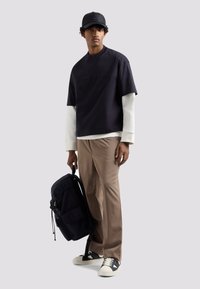 Top corto nero a maniche corte con un sottogiacca bianco, pantaloni beige, in spalla uno zaino scuro con cordoncino e indossando sneakers bianche e nere.