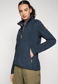 Fleece jacket in dark blue, featuring a high collar, zip front, and two side pockets with wooden accents. Worn over a plaid shirt.