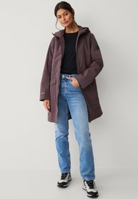Lange, met een capuchon voorziene jas in diep paars met een ritssluiting, twee voorzakken en een relaxte pasvorm. Gecombineerd met lichtblauwe jeans en zwarte sneakers.