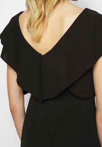 Femme aux cheveux blonds portant une robe noire avec un dos en V profond et des détails à volants sur les épaules et le haut du dos.