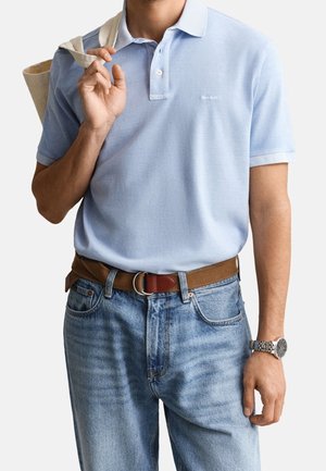 Homme portant un polo bleu clair, un jean bleu clair avec ceinture marron, une montre argentée au poignet et portant un sac fourre-tout beige sur l'épaule.