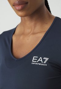Tengerészkék sportpóló V-nyakú kialakítással, sima anyagból készült. Az elején fehér EA7 Emporio Armani logó található.