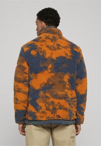 Giacca in pile con motivo astratto arancione e blu navy, collo alto, tessuto strutturato e orlo inferiore grigio. Chiusura con mezza zip.