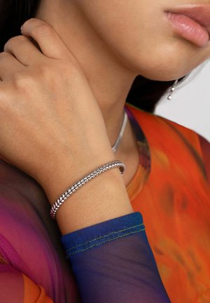 Bracciale in argento con design testurizzato indossato su un polso, abbinato a un top trasparente arancione e viola con un polsino blu.