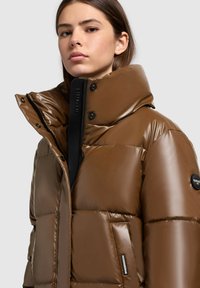 Jachetă căptușită maronie cu o finisare lucioasă, guler înalt, fermoar negru, nasturi cu apăsare și patch cu logo pe mânecă. Textură moale.