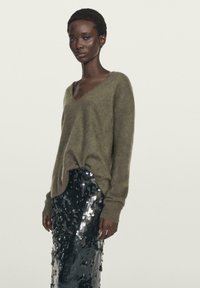 Woman wearing an olive green fuzzy V-neck sweater and a high-waisted black sequin skirt against a plain light background.