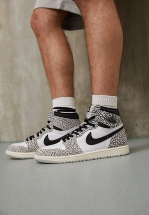 Jambes portant des baskets montantes Nike blanches et grises avec un swoosh noir et des détails à motifs, assorties à des chaussettes blanches mi-mollet.