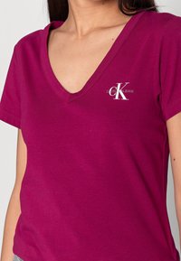 Magenta Baumwoll-T-Shirt mit V-Ausschnitt, kurzen Ärmeln und einem weißen Calvin Klein Jeans-Logo im linken Brustbereich. Sanfte Textur.