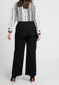 Pantalons noirs à jambes larges avec une texture lisse, dotés d'une taille haute et de plis sur les côtés, portés avec un haut à rayures à manches longues.