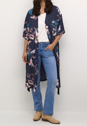Kobieta ubrana w jasnoniebieskie dżinsy, beżowe buty, białą koszulę i granatowe kwiatowe kimono, stoi z jedną ręką w kieszeni.