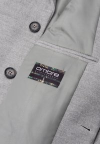 Interno di un cappotto grigio con una finitura di tessuto liscia, bottone nero e un'etichetta verde e nera con la dicitura "OMBRE QUINTESSENCE S."