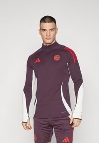 Maglietta sportiva a maniche lunghe color bordeaux con dettagli bianchi, zip a quarti e strisce rosse. Caratterizzata da un logo circolare sul petto e un design aderente.