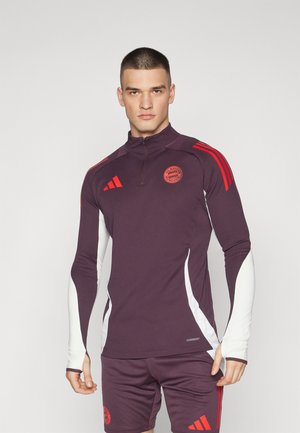 Maglietta sportiva a maniche lunghe color bordeaux con dettagli bianchi, zip a quarti e strisce rosse. Caratterizzata da un logo circolare sul petto e un design aderente.
