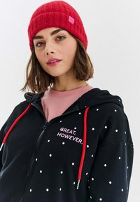 Czarna bluza z kapturem na zamek błyskawiczny w białe kropki, z czerwonymi sznurkami i różowym haftem "WSPANIE. JEDNAK.", założona na różową bluzkę, z czerwoną wełnianą czapką.