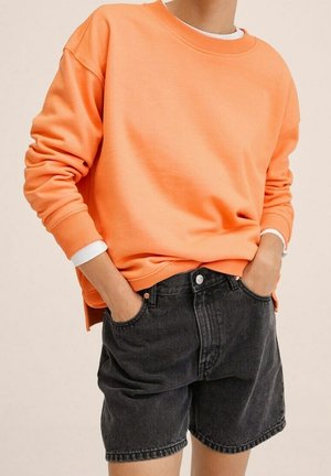 Personne portant un sweat-shirt orange ample par-dessus une chemise blanche, associé à un short en jean noir délavé taille haute, mains dans les poches.