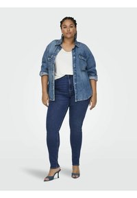 Camisa de mezclilla abotonada, de color azul claro; camiseta blanca debajo; vaqueros de tiro alto de color azul oscuro; sandalias de tacón azules; fondo neutral.