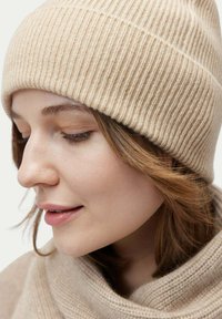 Beige ribbat stickad mössa, mjuk textur, snug passform. Modellen bär en matchande beige stickad scarf som smälter sömlöst in med hattens design.