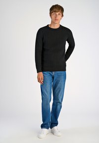 Maglione nero a maglia testurizzata con scollo a girocollo, abbinato a jeans azzurri chiari e sneakers bianche, in piedi su uno sfondo neutro.