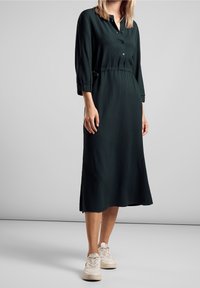 Abito midi verde scuro con una placca di bottoni, maniche a tre quarti, vita elastica e tessuto leggermente strutturato. Abbinato a sneakers bianche.