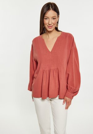 Blusa color rosa con scollatura a V profonda, vita arricciata e maniche lunghe e ampie. Tessuto morbido con una leggera lucentezza. Abbinata a pantaloni bianchi.